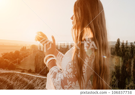 Happy woman in white boho dress on sunset in mountains. Romantic woman with long hair standing with her back on the sunset in nature in summer with open hands. Silhouette. Nature. Sunset. Happy woman in white boho dress on sunset in mountains. Romantic woman with long hair standing with her back on the sunset in nature in summer with open hands. Silhouette. Nature. Sunset. 106837738