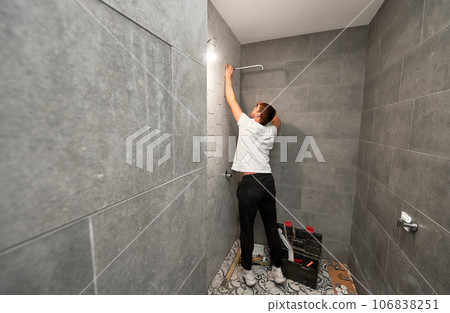 Male plumber fixing bathroom showerhead while working on bathroom renovation. Back view of man worker installing high pressure bath showerhead at home. Male plumber fixing bathroom showerhead while working on bathroom renovation. Back view of man worker installing high pressure bath showerhead at home. 106838251