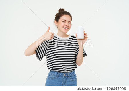 Portrait of beautiful, smiling brunette woman, shows thumbs up and bottle of vitamins, recommends dietary supplements, isolated against white background 106845008