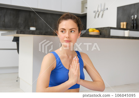 Young healthy beautiful woman in sports top and leggings practicing yoga at home, sitting in lotus position on yoga mat, meditating, smiling, relaxed, eyes closed, meditation concept 106845095