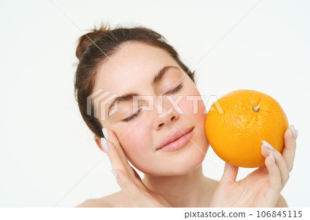 Portrait of young woman holding orange near face without blemishes, smiling, concept of vitamins, nourishment and organic facial treatments, white background 106845115