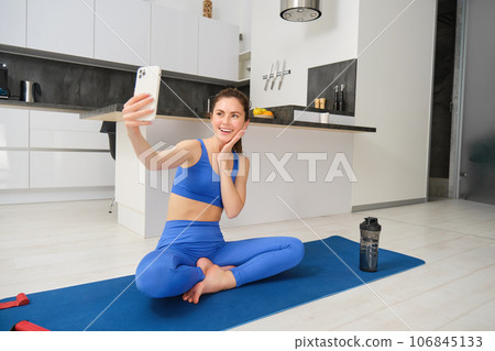 Young smiling woman, fitess girl doing workout from home, sits on yoga mat with smartphone, taking selfie, records sport vlog indoors Young smiling woman, fitess girl doing workout from home, sits on yoga mat with smartphone, taking selfie, records sport vlog indoors 106845133