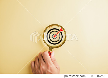A target board captured inside a magnifier glass focuses on business objectives against a backdrop with copy space. The image represents the concept of search, goal setting, strategy, and success. 106846258