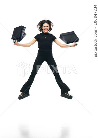 Portrait of happy, cheerful woman wearing fashion total black outfit holding bags in hands and jumping isolated on white studio color background. ad 106847234