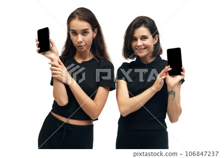 Portrait of two young happy women wearing fashion outfit holding phones with blank screen in hands isolated on white studio color background. ad Portrait of two young happy women wearing fashion outfit holding phones with blank screen in hands isolated on white studio color background. ad 106847237