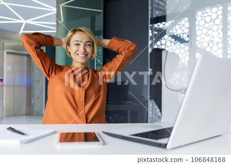 Well done woman completed work, businesswoman at workplace inside office smiling and looking at camera, female worker threw hands behind head satisfied with achievement results. 106848168