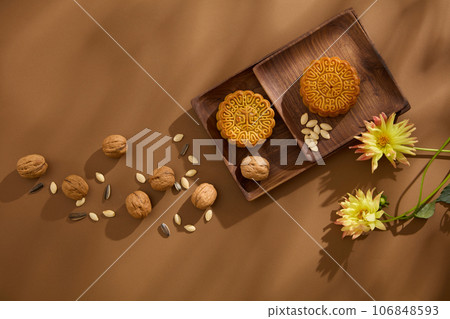 Wooden antique dishes with tasty mooncakes placed on. Walnuts, sunflower seeds and pumpkin seeds are scattered over brown background with flowers. Happy Mid Autumn Festival Wooden antique dishes with tasty mooncakes placed on. Walnuts, sunflower seeds and pumpkin seeds are scattered over brown background with flowers. Happy Mid Autumn Festival 106848593