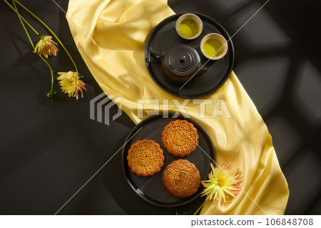 Top view of mooncakes displayed on round dishes with tea set, yellow flower and yellow silk cloth on black background. Scene for advertising. Traditional Chinese mid-autumn festival 106848708