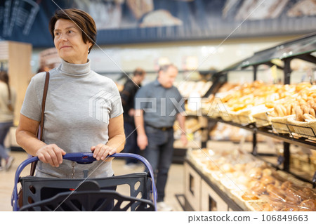 Elderly female shopper with a shopping cart chooses groceries in supermarket 106849663