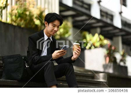 Handsome businessman in formal suit using smartphone, sitting on the stairs of building Handsome businessman in formal suit using smartphone, sitting on the stairs of building 106850436
