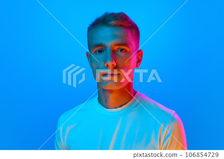 Portrait of young good-natured, calm man, student in white t-shirt looking at camera isolated on blue background in red neon light. 106854729