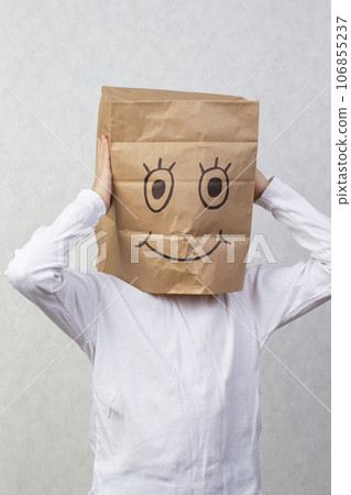 The boy has a cardboard box above his head with a funny smiley face drawn on it. 106855237