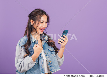 Portrait Asian young woman confident wear denim jeans shirt smiling excited using smart mobile phone, female surprised making winner gesture on smartphone, studio shot isolated on purple background 106856801