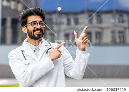 A young Hindu doctor from outside the clinic smiles and looks away, the man points his fingers in the direction, smiles with satisfaction and the doctor with a medical coat and a stethoscope. A young Hindu doctor from outside the clinic smiles and looks away, the man points his fingers in the direction, smiles with satisfaction and the doctor with a medical coat and a stethoscope. 106857329