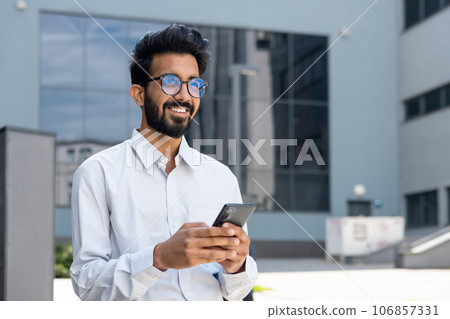 A young man, a businessman, a programmer walks through the city, holds the phone in his hands, smiles contentedly, types messages and browses the Internet online. A young man, a businessman, a programmer walks through the city, holds the phone in his hands, smiles contentedly, types messages and browses the Internet online. 106857331