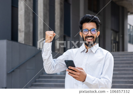 Arab online winner portrait, man in business shirt walking around the city outside office building, businessman smiling and looking at camera, holding hand up triumph gesture, using app on phone. Arab online winner portrait, man in business shirt walking around the city outside office building, businessman smiling and looking at camera, holding hand up triumph gesture, using app on phone. 106857332