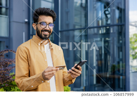 Portrait of young man outside office building, hispanic smiling and looking at camera, holding phone and bank credit card, online shopping and booking services remotely. Portrait of young man outside office building, hispanic smiling and looking at camera, holding phone and bank credit card, online shopping and booking services remotely. 106857999