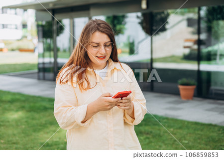 Happy cheerful young woman walking on city street checks her smartphone. Portrait of beautiful 30s businesswoman using smartphone outdoors. Urban lifestyle concept. Happy cheerful young woman walking on city street checks her smartphone. Portrait of beautiful 30s businesswoman using smartphone outdoors. Urban lifestyle concept. 106859853