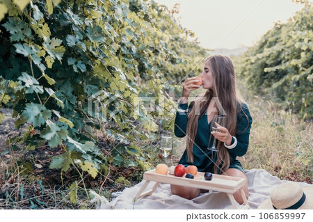Woman picnic vineyard. Happy woman with a glass of wine at a picnic in the vineyard, wine tasting at sunset and open nature in the summer. Romantic dinner, fruit and wine. Woman picnic vineyard. Happy woman with a glass of wine at a picnic in the vineyard, wine tasting at sunset and open nature in the summer. Romantic dinner, fruit and wine. 106859997
