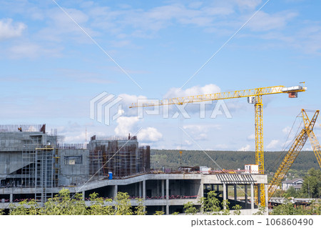 A crane and a building under construction against a blue sky background. Builders work on large construction sites, and there are many cranes working in the field of new construction. A crane and a building under construction against a blue sky background. Builders work on large construction sites, and there are many cranes working in the field of new construction. 106860490