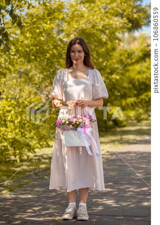 Beautiful girl with flowers, in a dress, glasses and sneakers. A beautiful bouquet of flowers in a box in the hands of a beautiful girl who walks along the street on a sunny day. Beautiful girl with flowers, in a dress, glasses and sneakers. A beautiful bouquet of flowers in a box in the hands of a beautiful girl who walks along the street on a sunny day. 106860559