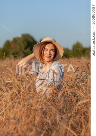 A young beautiful girl with braces on her teeth and long hair poses in a wheat field in the summer at sunset. The girl holds a hat in her hand against the background of a wheat field. A young beautiful girl with braces on her teeth and long hair poses in a wheat field in the summer at sunset. The girl holds a hat in her hand against the background of a wheat field. 106860587