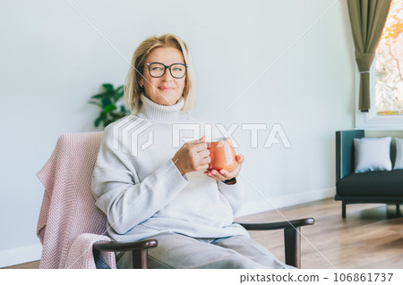 Portrait of smiling middle aged woman relaxing with cup of hot drink in scandy style hygge modern interior home. Lady dreaming, enjoy calm mood without stress, well being alone. Cozy autumn 106861737