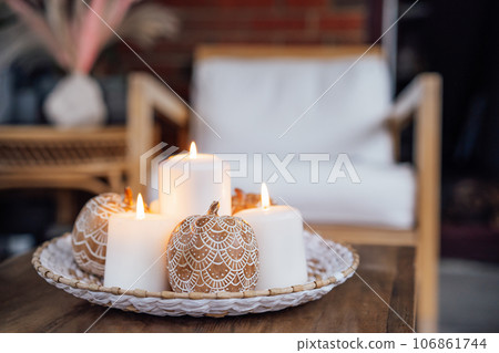 Autumn fall cozy mood composition for hygge home decor. Orange pumpkins decorated with mandalas, white burning candles on wicker tray on the coffee table in the loft living room. Selective focus 106861744