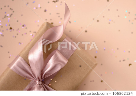 Xmas gift package in kraft paper with pink ribbon on trendy natural beige background with glitter. Zero waste Christmas presents packaging. Happy holidays celebration concept. 2024, 2025 flat lay 106862124