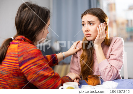 Woman comforting her friend while sitting at table with cups of tea Woman comforting her friend while sitting at table with cups of tea 106867245