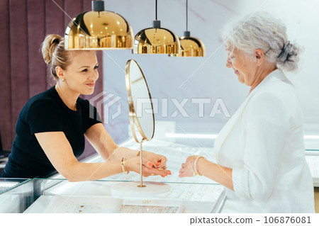 Elderly grey haired woman buying some jewelry. Woman owner, saleswoman of jewelry shop presenting for senior female consumer golden ring with diamond. 106876081