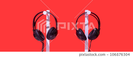 Two pairs of black headphones on stand. Red background, white table and black noise reduction device create creative atmosphere. Individual lecture, listening music, audio training, concern for others 106876304