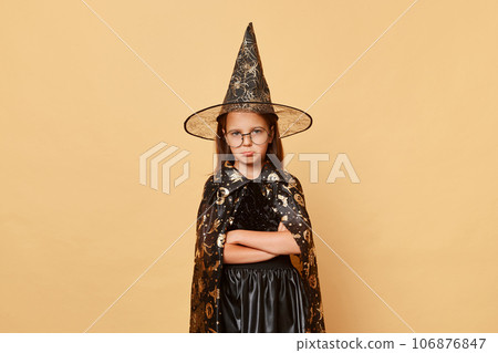 Sad offended female kid girl wearing witch black hat costume isolated over beige background expressing sorrow and sadness keeps hands crossed being bored at halloween party. Sad offended female kid girl wearing witch black hat costume isolated over beige background expressing sorrow and sadness keeps hands crossed being bored at halloween party. 106876847