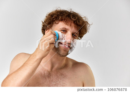 Shirtless mature man with curly hair taking care after skin, washing face with foam and brush against grey studio background Shirtless mature man with curly hair taking care after skin, washing face with foam and brush against grey studio background 106876878