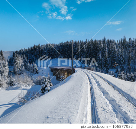 Stone viaduct (arch bridge) on railway through mountain snowy fir forest. Snow drifts on wayside and hoarfrost on trees and electric line wires. Stone viaduct (arch bridge) on railway through mountain snowy fir forest. Snow drifts on wayside and hoarfrost on trees and electric line wires. 106877083