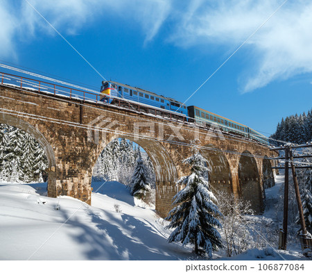 Stone viaduct (arch bridge) on railway through mountain snowy fir forest and locomotive with a passenger train. Snow drifts on wayside and hoarfrost on trees and electric line wires. Stone viaduct (arch bridge) on railway through mountain snowy fir forest and locomotive with a passenger train. Snow drifts on wayside and hoarfrost on trees and electric line wires. 106877084