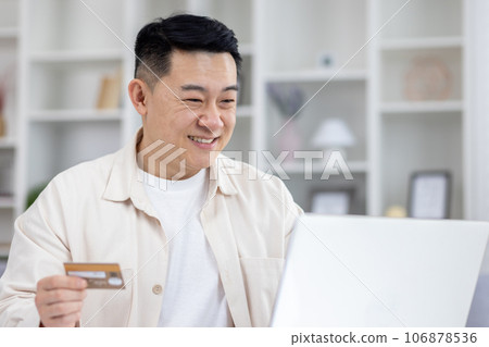 Joyful successful asian businessman working from bright home office, man using bank credit card and laptop for online shopping in online store, and booking services remotely. 106878536