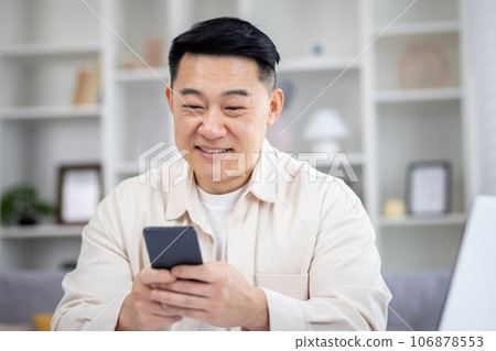 Mature Asian man sitting at table at home, man using phone, typing message on smartphone, app user and browsing internet in home cinema room, smiling contentedly. Mature Asian man sitting at table at home, man using phone, typing message on smartphone, app user and browsing internet in home cinema room, smiling contentedly. 106878553