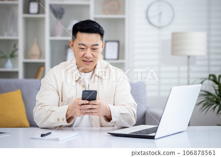Mature Asian man sitting at table at home, man using phone, typing message on smartphone, app user and browsing internet in home cinema room, smiling contentedly. Mature Asian man sitting at table at home, man using phone, typing message on smartphone, app user and browsing internet in home cinema room, smiling contentedly. 106878561