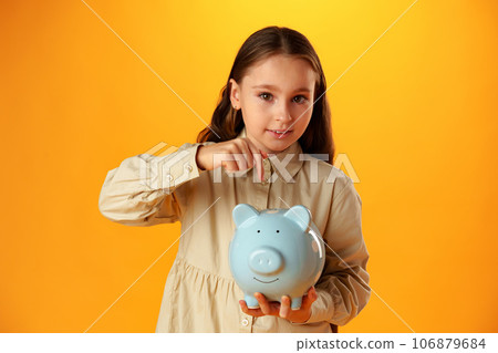 Smiling little girl posing with a piggy bank over yellow background Smiling little girl posing with a piggy bank over yellow background 106879684
