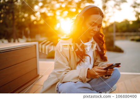 Young woman in headphones listening music, chatting outdoors. Lifestyle, relax, technology concept. Young woman in headphones listening music, chatting outdoors. Lifestyle, relax, technology concept. 106880400