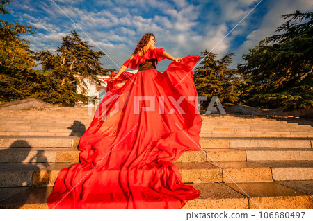 Sunrise red dress. A woman in a long red dress against the backdrop of sunrise, bright golden light of the sun's rays. The concept of femininity, harmony. 106880497