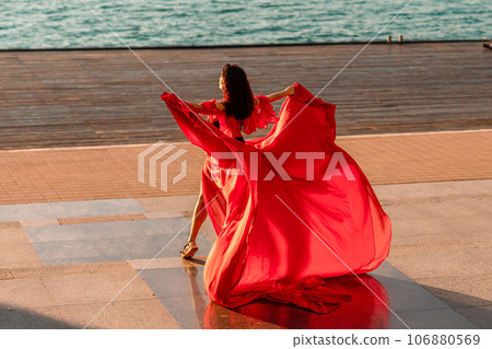 Sunrise red dress. A woman in a long red dress against the backdrop of sunrise, bright golden light of the sun's rays. The concept of femininity, harmony. 106880569