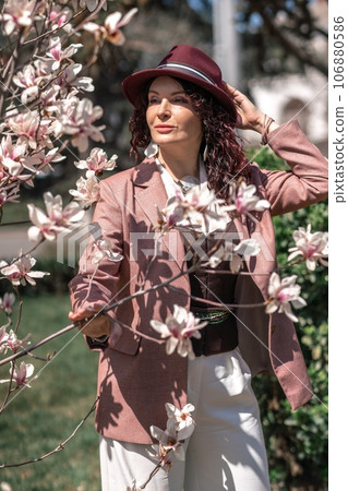 Magnolia park woman. Stylish woman in a hat stands near the magnolia bush in the park. Dressed in white corset pants and posing for the camera. Magnolia park woman. Stylish woman in a hat stands near the magnolia bush in the park. Dressed in white corset pants and posing for the camera. 106880586
