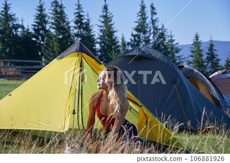 Woman traveller camping outdoors. Attractive, young female traveling, hiking in mountains. Sporty, slim woman sitting on grass near tent, smiling, admiring landscape in morning. Woman traveller camping outdoors. Attractive, young female traveling, hiking in mountains. Sporty, slim woman sitting on grass near tent, smiling, admiring landscape in morning. 106881926