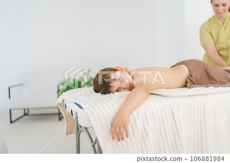 Cropped shot of professional female masseuse giving therapeutic massage of calf muscles to five year old boy lying on table in spa salon. Little kid getting feet massage treatment, looking at camera. 106881984