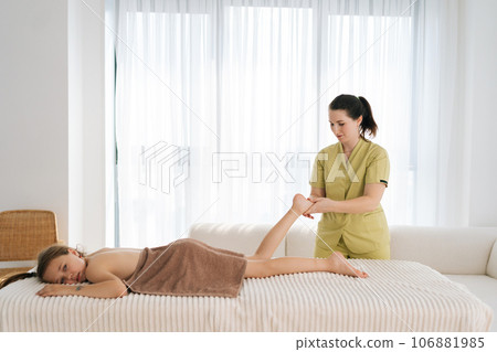 Side view portrait of female masseuse doing foot and legs massage to adorable ten-year-old girl lying on massage table in spa salon. Masseur kneads foot and leg of little kid by window. 106881985