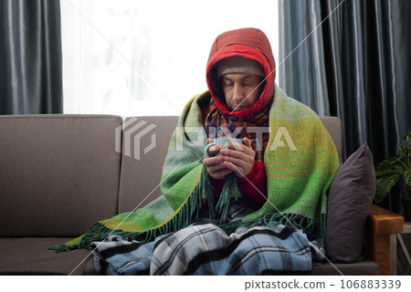 Middle age male model in hat parka and plaid hold coffee cup sitting on the sofa in his cold living room 106883339