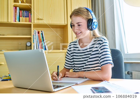 Girl teen student in headphones sitting at desk using computer writing in study notebook 106884019