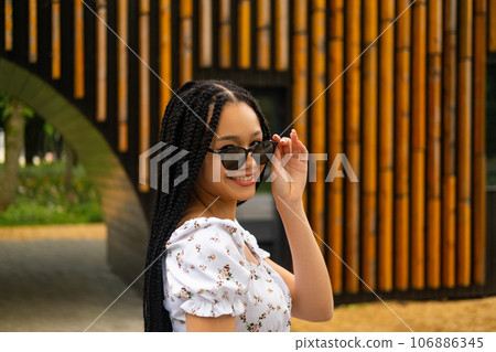Young African beautiful woman holding dark sunglasses with her fingers smiling and looking at the camera on the street against the background of a wooden wall in the park Young African beautiful woman holding dark sunglasses with her fingers smiling and looking at the camera on the street against the background of a wooden wall in the park 106886345